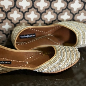 May include: A pair of gold and silver embellished flat shoes with a pointed toe. The shoes have a leather sole and are decorated with rows of sequins and beads.