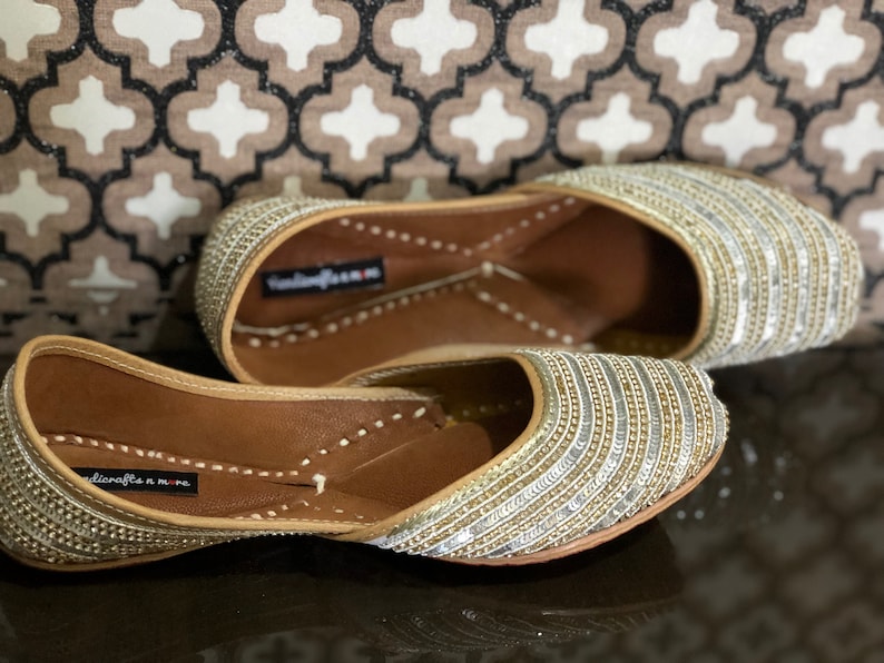 May include: A pair of gold and silver embellished flat shoes with a pointed toe. The shoes have a leather sole and are decorated with rows of sequins and beads.