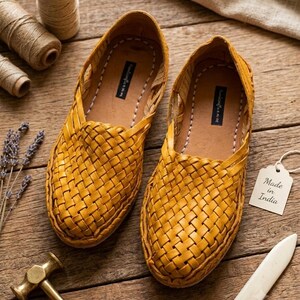 May include: A pair of mustard yellow woven leather slip-on shoes featuring a closed-toe design with a braided upper pattern. The interior insoles have a black label with white text. A white tag attached to the shoe reads Made in India.