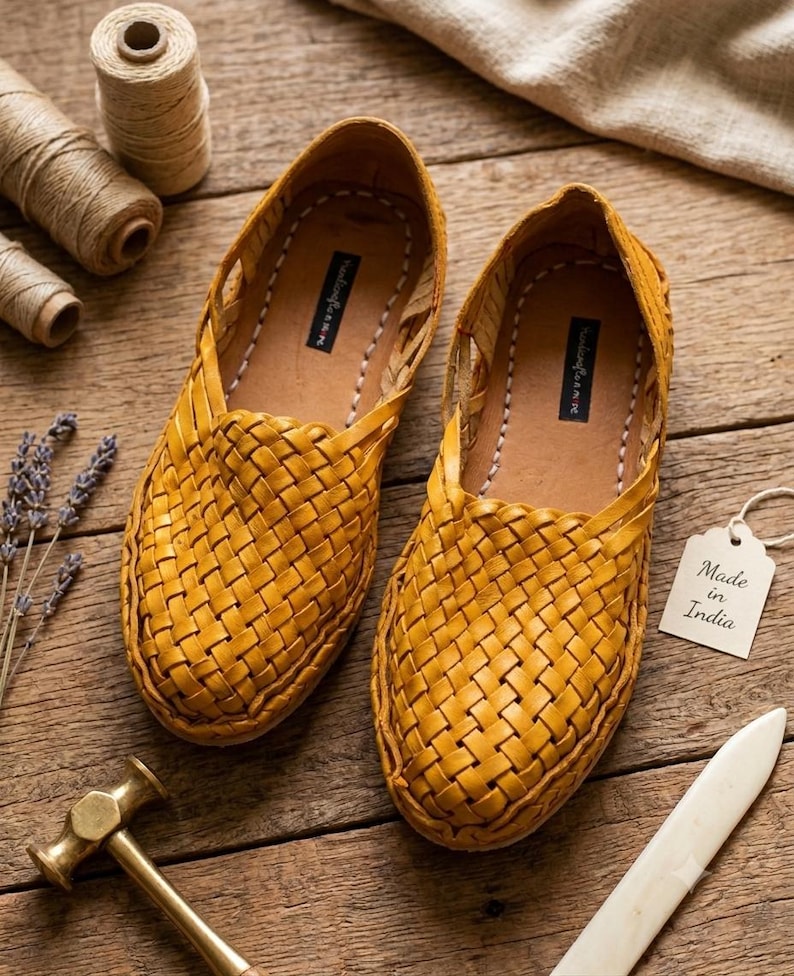 May include: A pair of mustard yellow woven leather slip-on shoes featuring a closed-toe design with a braided upper pattern. The interior insoles have a black label with white text. A white tag attached to the shoe reads Made in India.
