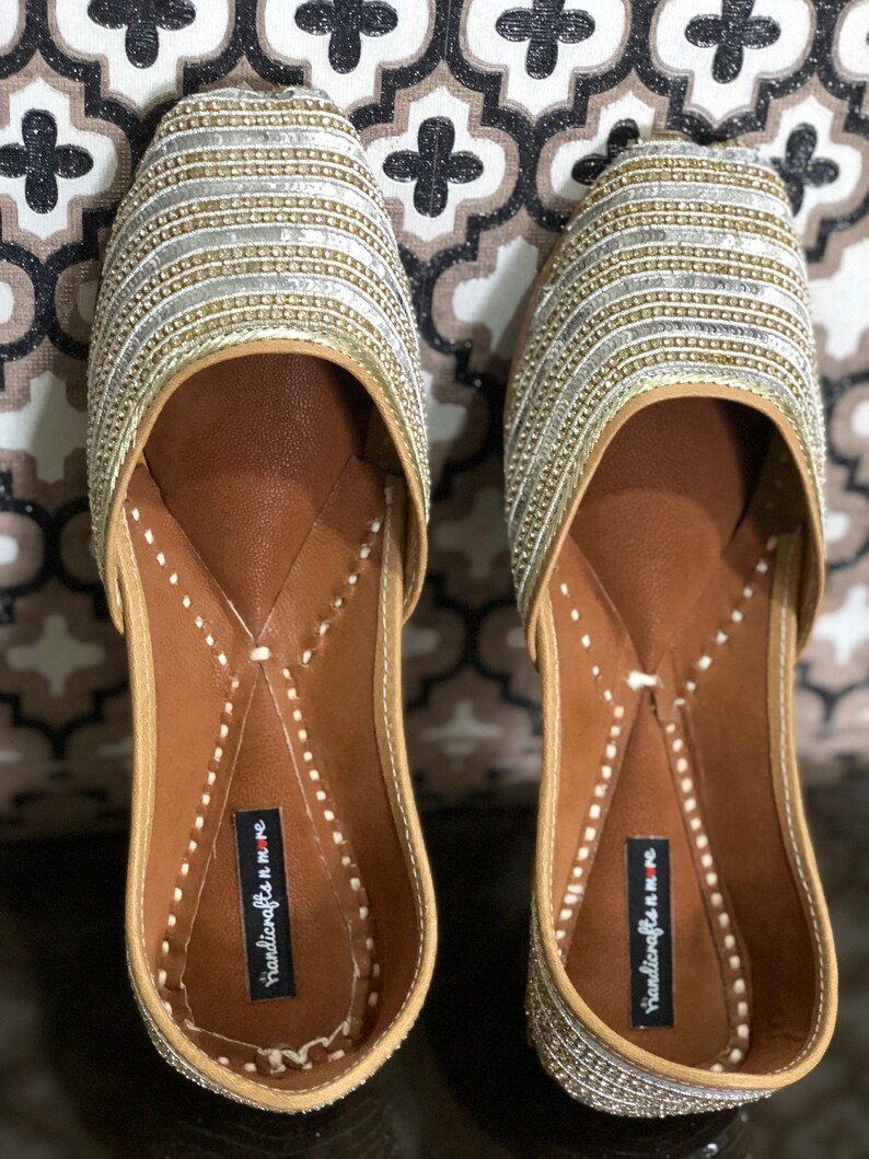 May include: Pair of gold and silver embellished flat shoes with a pointed toe and a brown leather insole. The shoes have a decorative stitching detail around the edge of the insole. The insole is labeled "handicrafts n more".