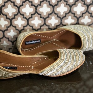 May include: A pair of gold and silver embellished flat shoes with a pointed toe. The shoes have a leather sole and are decorated with rows of sequins and beads.