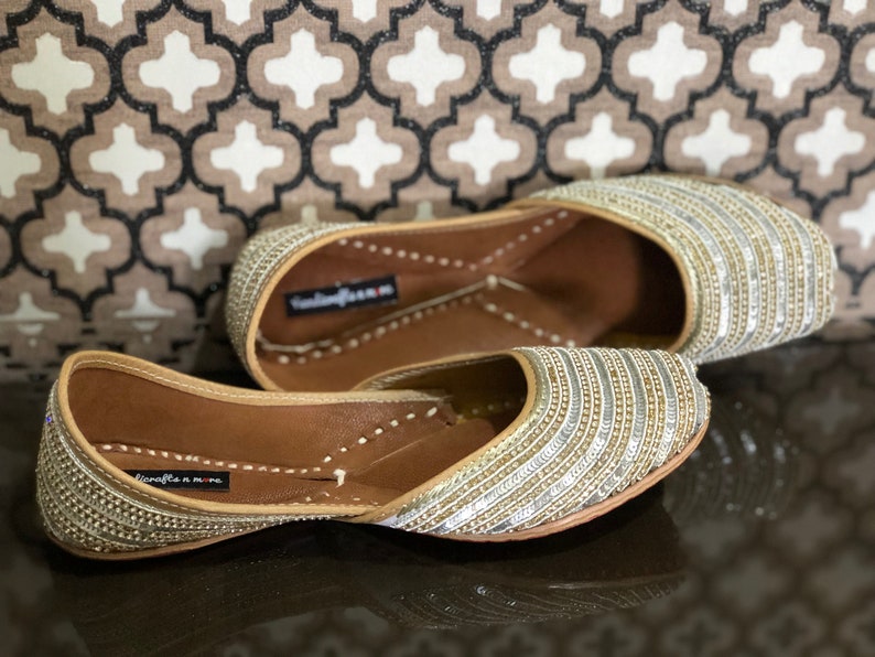 May include: A pair of gold and silver embellished flat shoes with a pointed toe. The shoes have a leather sole and are decorated with rows of sequins and beads.