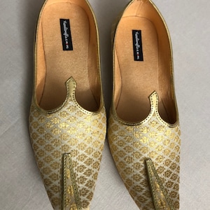 May include: A pair of gold and white patterned flat shoes with pointed toes. The shoes have a simple design and are made of a soft fabric. The brand name "Handicrafts n more" is printed on the inside of the shoes.