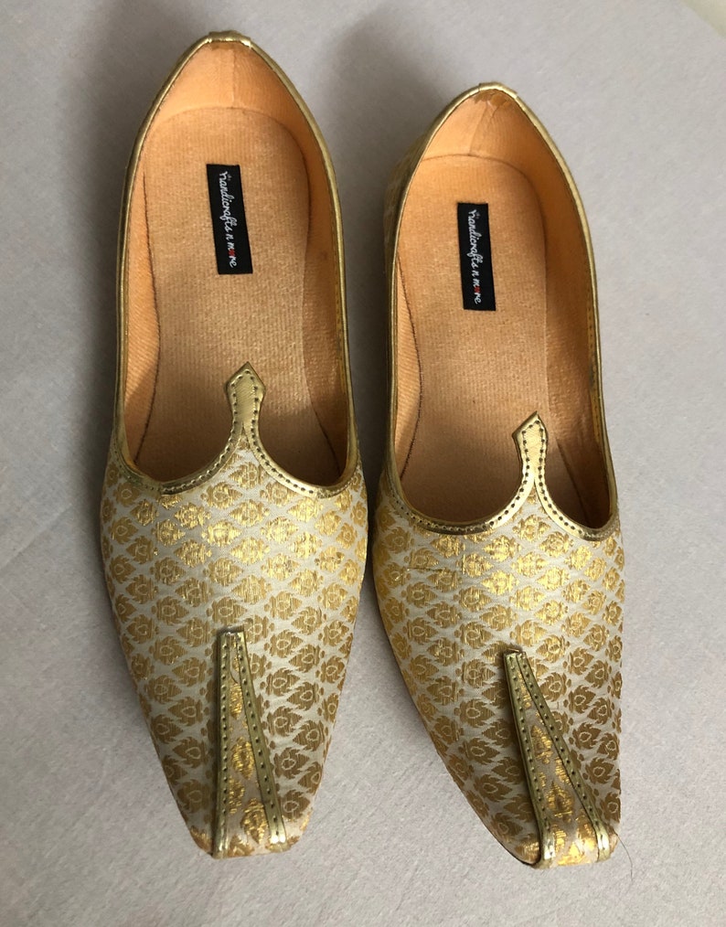 May include: A pair of gold and white patterned flat shoes with pointed toes. The shoes have a simple design and are made of a soft fabric. The brand name "Handicrafts n more" is printed on the inside of the shoes.