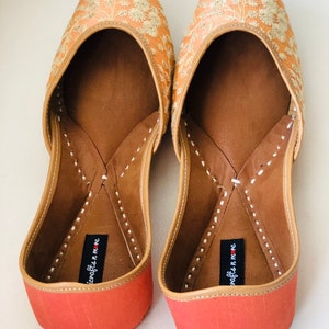 May include: A pair of peach-coloured embroidered flat shoes with a red sole and the text "juxtapose n me" on the inside of each shoe.