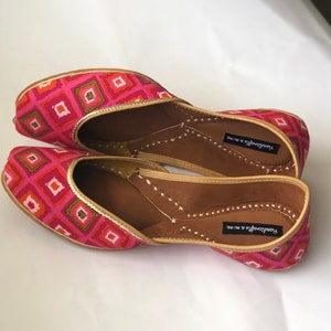 May include: A pair of pink and gold patterned flat shoes with a pointed toe and a small opening at the top. The shoes have a brown leather insole and are labeled "Yandavarga & More".