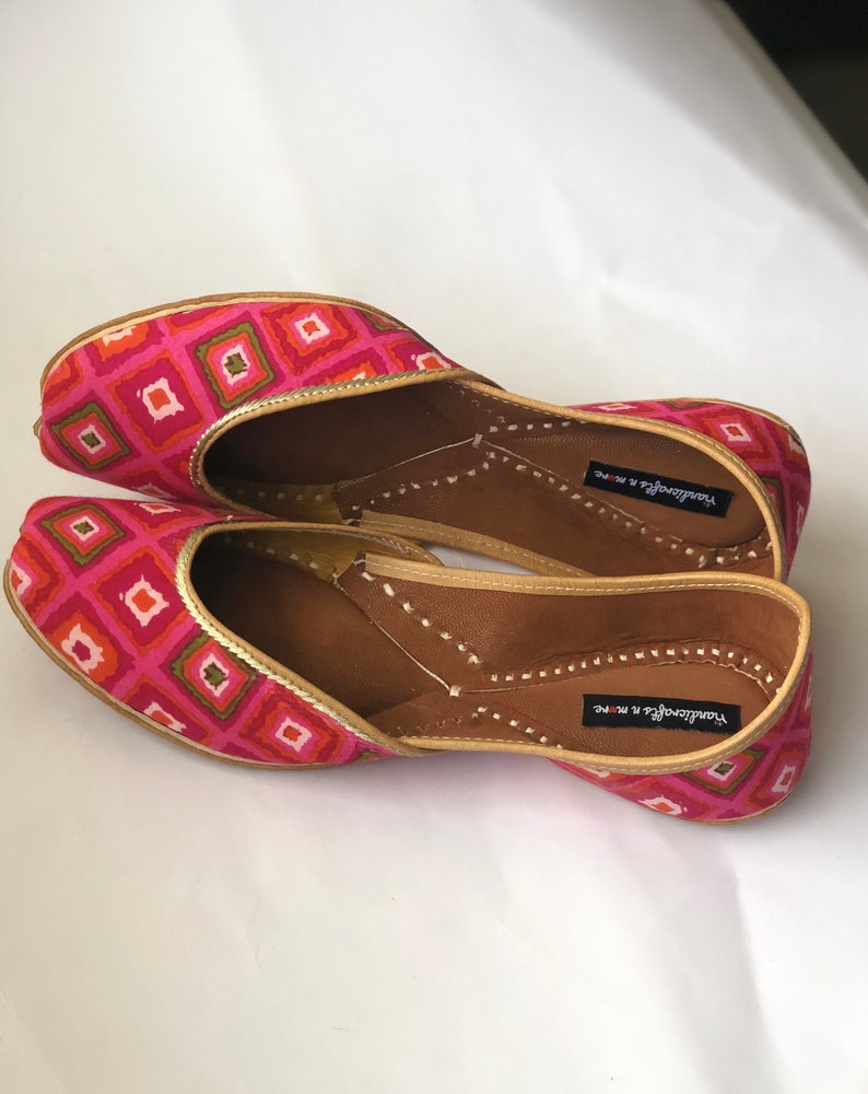 May include: A pair of pink and gold patterned flat shoes with a pointed toe and a small opening at the top. The shoes have a brown leather insole and are labeled "Yandavarga & More".