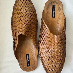 May include: A pair of brown leather woven slip-on sandals. The sandals have a textured, basket-weave design on the top and a smooth leather insole. The brand name "handicrafts n.m.re" is printed on the insole.