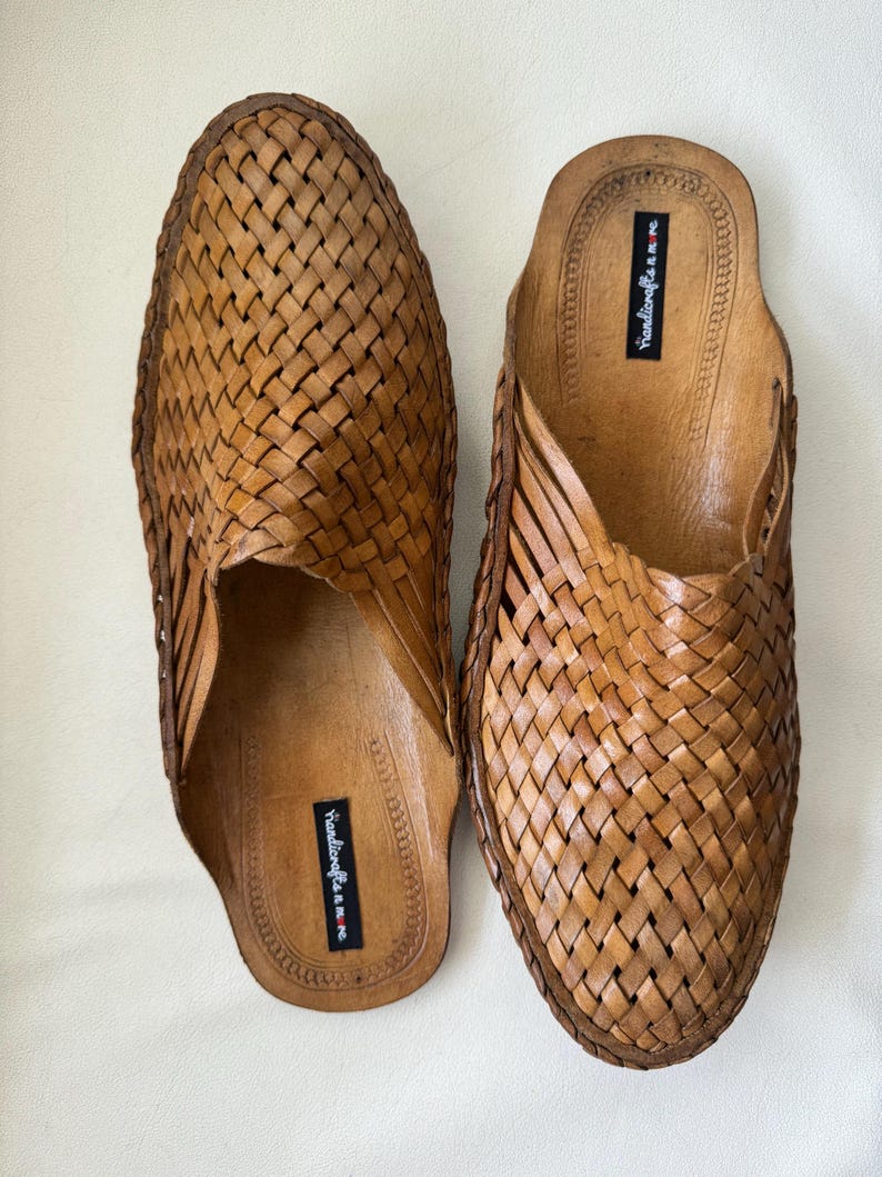 May include: A pair of brown leather woven slip-on sandals. The sandals have a textured, basket-weave design on the top and a smooth leather insole. The brand name "handicrafts n.m.re" is printed on the insole.