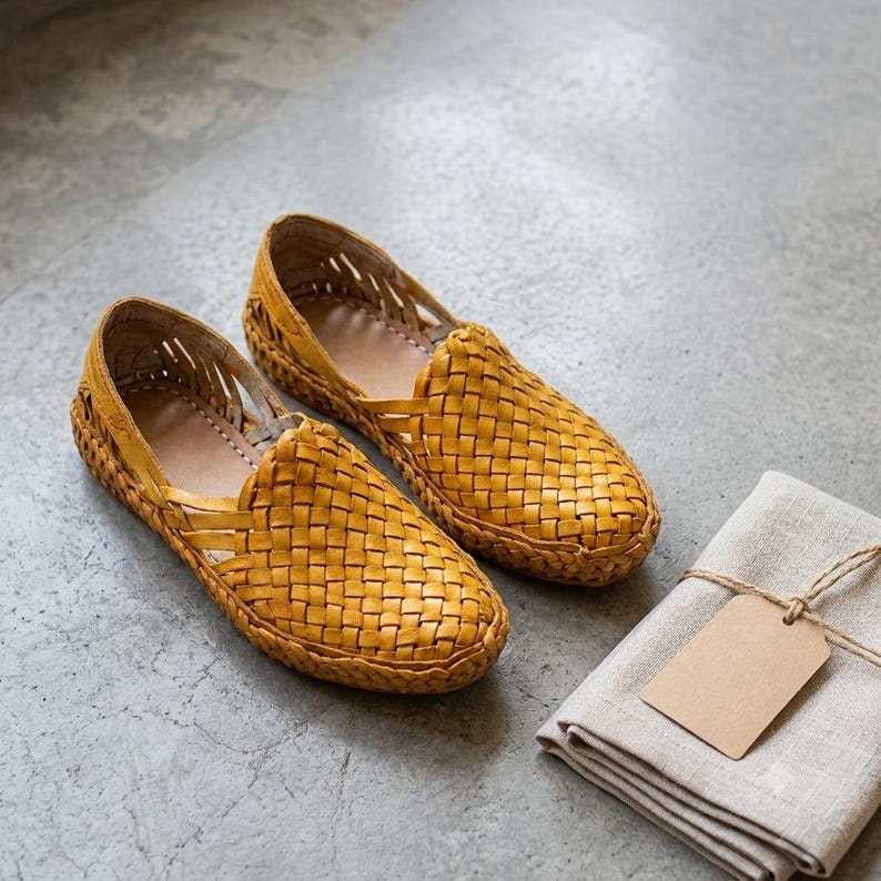 May include: A pair of mustard yellow slip-on shoes featuring a woven leather upper with a lattice design on the sides and a rounded toe. Beside the shoes lies a folded light beige linen cloth with a blank rectangular tan paper tag attached.