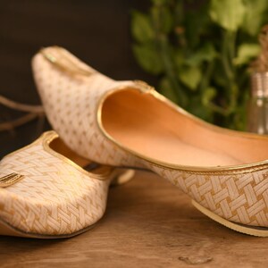 May include: A pair of white and gold patterned flat shoes with pointed toes. The shoes have a woven design and a gold trim.