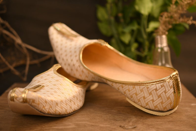 May include: A pair of white and gold patterned flat shoes with pointed toes. The shoes have a woven design and a gold trim.