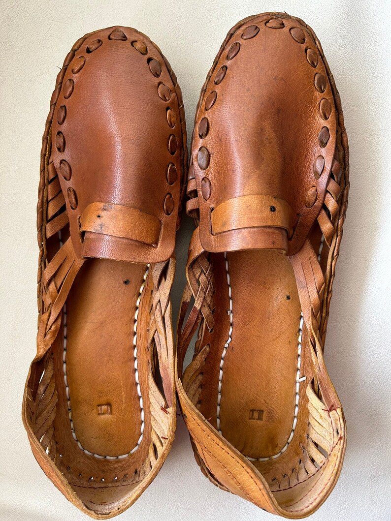 May include: A pair of tan leather huarache-style slip-on shoes featuring a woven side construction, a solid leather vamp with a decorative strap, and a stitched border detail. The interior footbed shows white stitching and a small stamped mark.