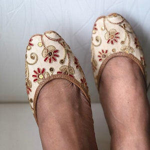 May include: A pair of cream-colored flat shoes with gold and red embroidery. The shoes have a pointed toe and a closed back.