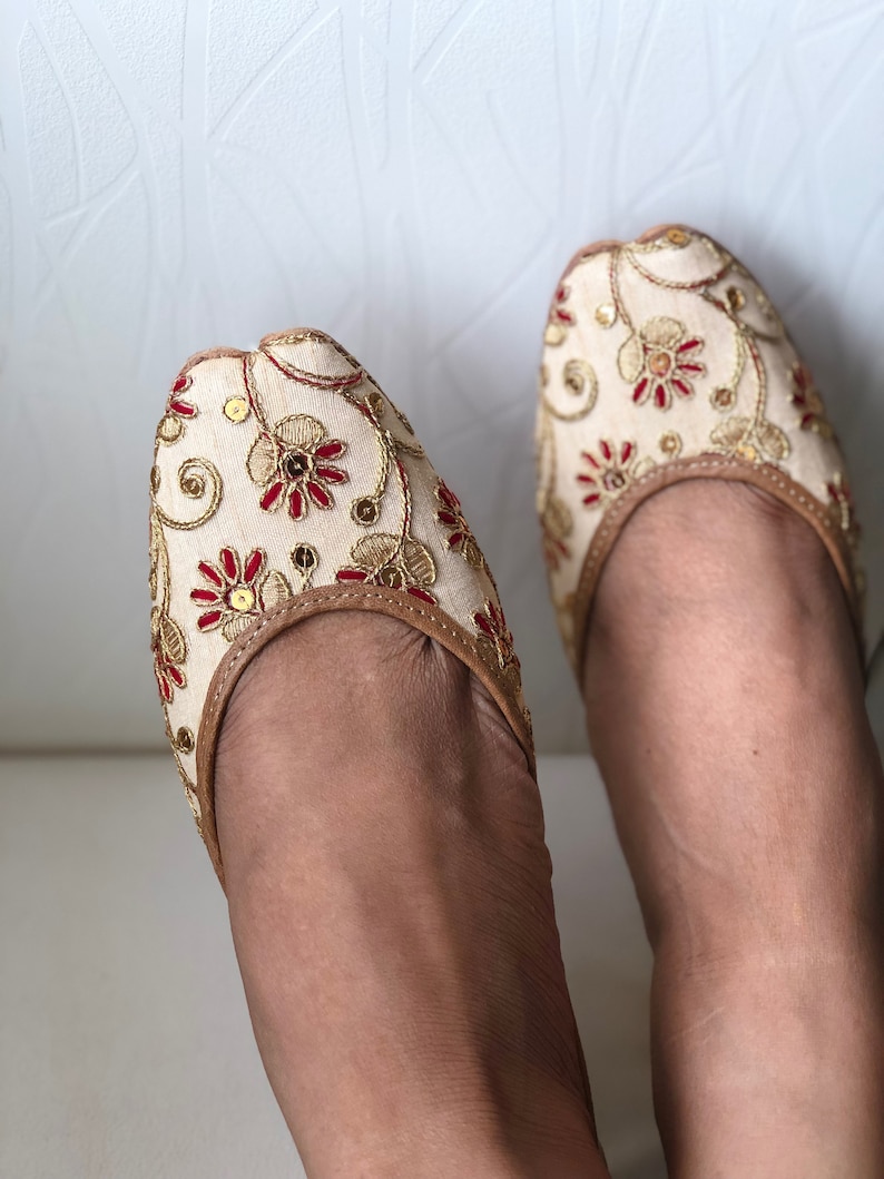 May include: A pair of cream-colored flat shoes with gold and red embroidery. The shoes have a pointed toe and a closed back.