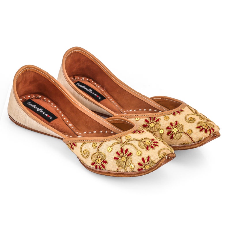 May include: A pair of beige embroidered flat shoes with red and gold floral designs. The shoes have a pointed toe and a low heel. The brand name "Handicrafts of India" is printed on the inside of the shoes.