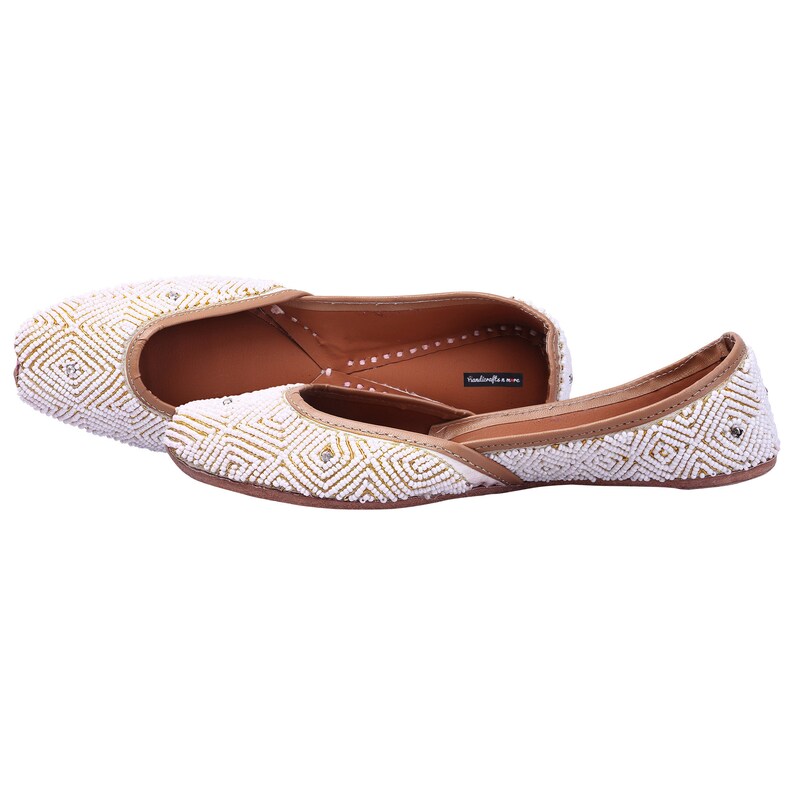 May include: A pair of white and gold embellished flat shoes with a geometric pattern. The shoes have a brown leather sole and a pointed toe.