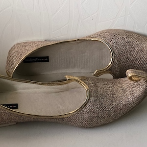 K&ouml;nnte beinhalten: Ein Paar goldene und braune Stoffschuhe mit spitzer Zehenpartie und einem dekorativen Detail an der Oberseite. Die Schuhe haben einen goldenen Rand.