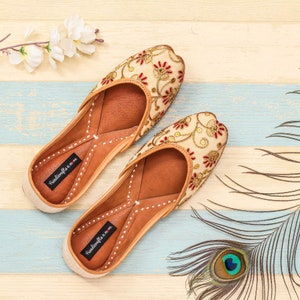 May include: A pair of cream-colored embroidered flat shoes with red and gold accents. The shoes have a pointed toe and a small heel. The brand name "Handicrafts n More" is printed on the insole of each shoe.
