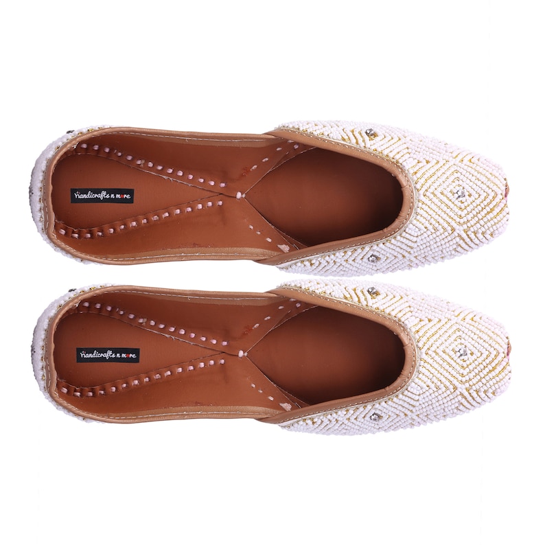 May include: A pair of white beaded flat shoes with gold accents. The shoes have a pointed toe and a low heel. The brand name "handicrafts n more" is visible on the insole.