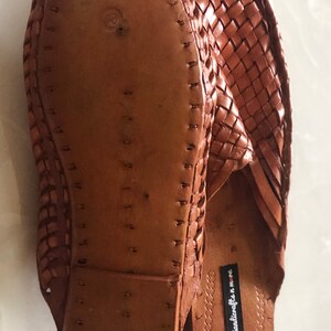 May include: A pair of brown leather woven slippers with a braided design. The soles are made of leather and have a stitched edge. The slippers have a label that reads "Handicrafts in MMC".