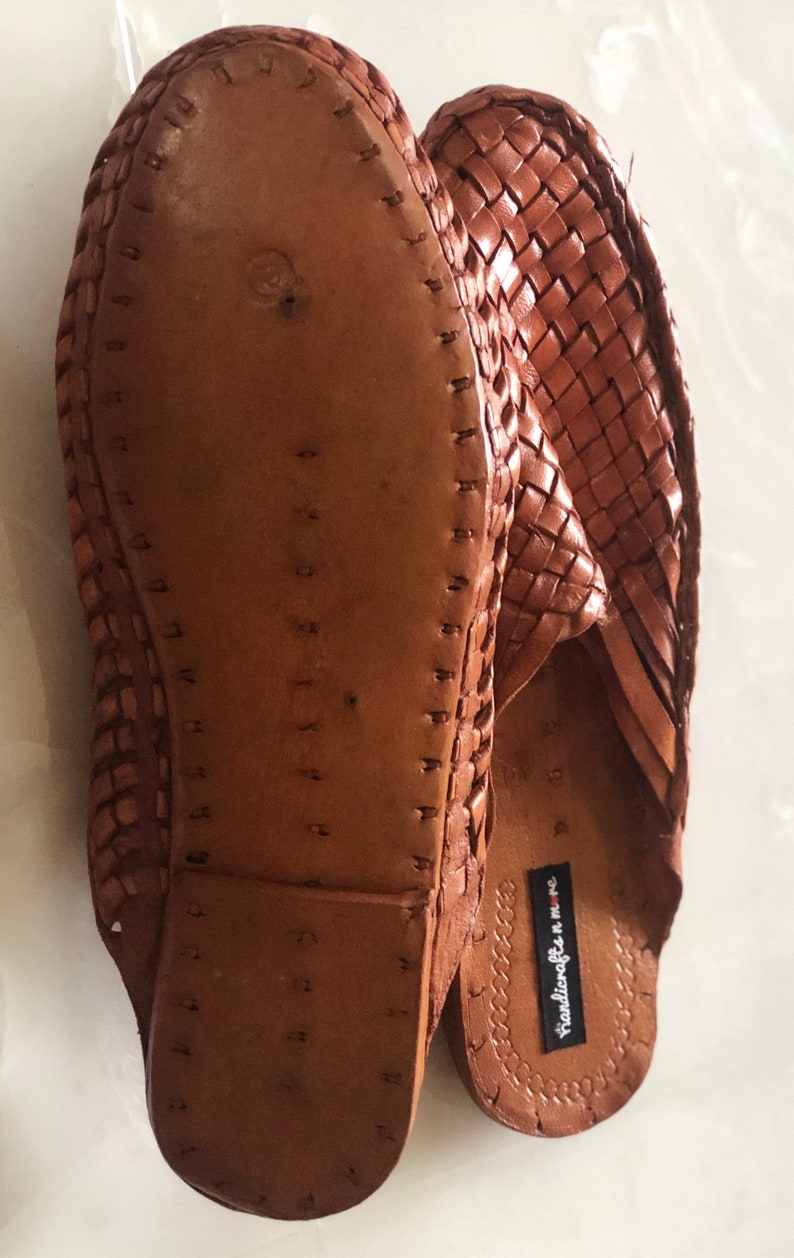 May include: A pair of brown leather woven slippers with a braided design. The soles are made of leather and have a stitched edge. The slippers have a label that reads "Handicrafts in MMC".