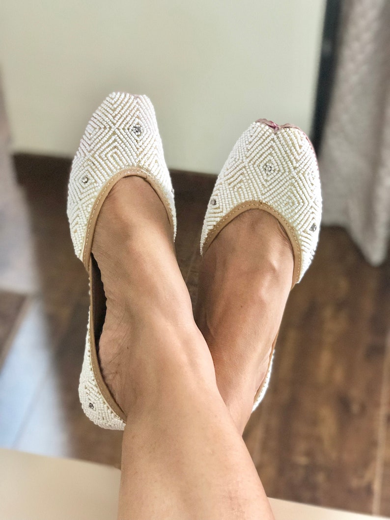 May include: White beaded flat shoes with a geometric pattern. The shoes have a pointed toe and a low heel.