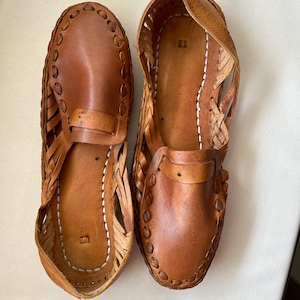 May include: A pair of tan leather huarache-style shoes featuring a woven side design, a solid toe cap, and a strap across the vamp. The interior soles are stitched with white thread and feature a small square embossed mark near the heel area.