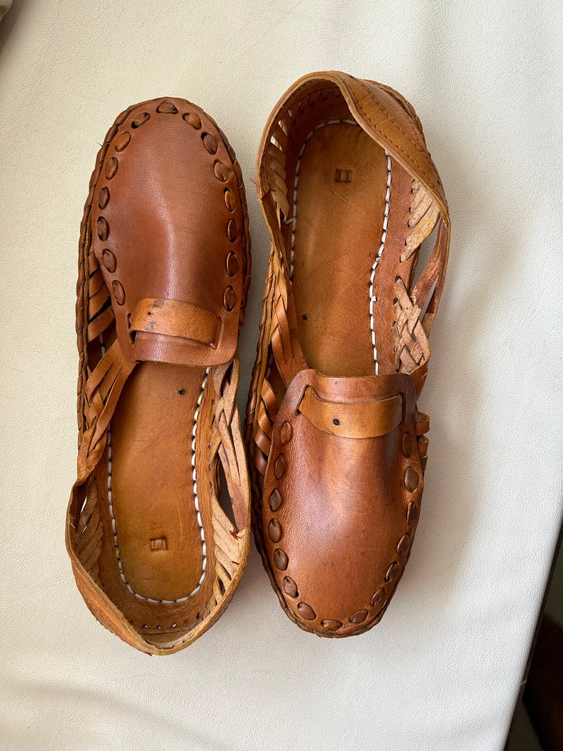 May include: A pair of tan leather huarache-style shoes featuring a woven side design, a solid toe cap, and a strap across the vamp. The interior soles are stitched with white thread and feature a small square embossed mark near the heel area.
