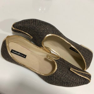 May include: A pair of brown woven flat shoes with gold trim. The shoes have a pointed toe and a closed back. The brand name "Handicrafts & More" is visible on the inside of one shoe.