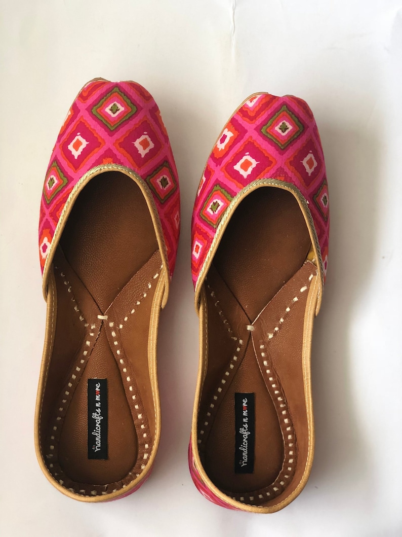 May include: A pair of pink and green flat shoes with a geometric pattern. The shoes have a gold trim and a brown leather insole. The brand name "handicrafts n more" is printed on the insole.