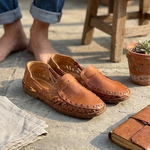 May include: A pair of tan leather loafers featuring a woven side design and a smooth front panel with a decorative stitched border. The shoes have a low profile and a flat sole, showcasing intricate braided leather detailing along the sides.