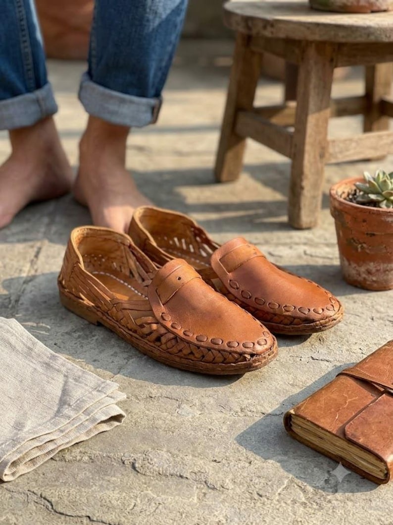 May include: A pair of tan leather loafers featuring a woven side design and a smooth front panel with a decorative stitched border. The shoes have a low profile and a flat sole, showcasing intricate braided leather detailing along the sides.