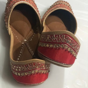 May include: A pair of red embroidered flat shoes with gold and silver embellishments. The shoes have a pointed toe and a low heel.