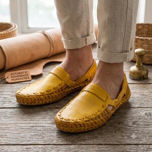May include: A pair of yellow leather huarache sandals featuring a woven design along the edges and a strap across the top. A small rectangular tag with the text AUTHENTIC HANDMADE HUARACHES is visible next to the footwear on a wooden surface.