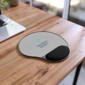 May include: An oval-shaped computer mouse pad with a gray surface and a black wrist rest. The pad has a silver trim and the words "TROPHY POINT" are printed on the surface in a teal color.
