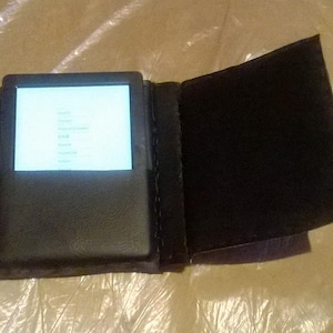 Puede incluir: Funda de lector electrónico de cuero negro, abierta para mostrar la pantalla. La pantalla está iluminada, mostrando una lista de opciones de idioma. La funda está abierta, revelando el forro interior. La funda está sobre una superficie arrugada y clara.
