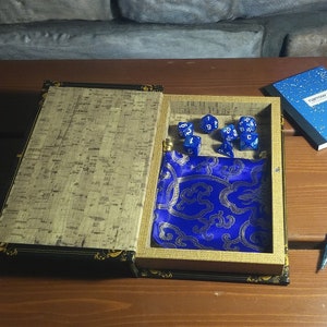 Könnte beinhalten: Ein blauer Samt-Würfelbeutel befindet sich in einer Holzkiste in Buchform mit einem korkartigen Innenraum. Die Kiste ist geöffnet und es befinden sich mehrere blaue Würfel mit weißen Zahlen in der Kiste.
