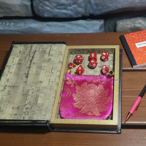 Könnte beinhalten: Ein offenes Buch mit einem Holzinnenraum, der einen rosa Samt-Würfelbeutel und rote Würfel mit weißen Zahlen enthält. Das Buch liegt auf einem Holztisch mit einem roten Notizbuch und einem rosa Stift.