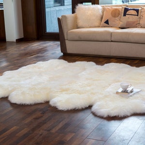 May include: A white sheepskin rug with a fluffy texture, laid out on a hardwood floor in a living room setting. A white couch with throw pillows is in the background.