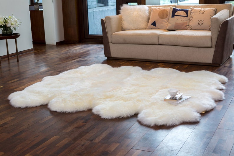 May include: A white sheepskin rug with a fluffy texture, laid out on a hardwood floor in a living room setting. A white couch with throw pillows is in the background.