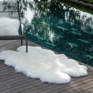 May include: A white sheepskin rug placed on a wooden deck next to a black metal chair with a white fluffy pillow. The rug is in front of a swimming pool.