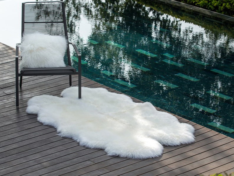 May include: A white sheepskin rug placed on a wooden deck next to a black metal chair with a white fluffy pillow. The rug is in front of a swimming pool.