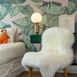 May include: A white faux fur chair with wooden legs sits in a room with a pink and green tropical leaf wallpaper. A white table with a green top holds a white globe lamp with a gold base and a small giraffe figurine.