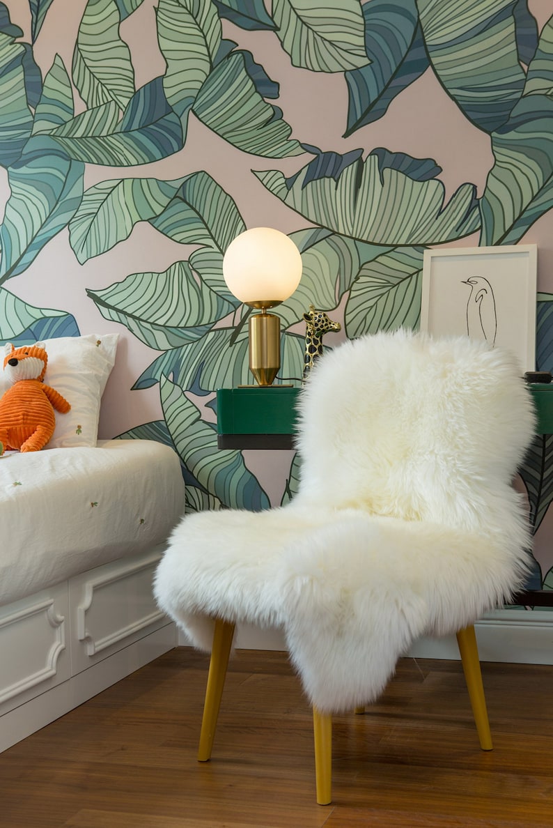 May include: A white faux fur chair with wooden legs sits in a room with a pink and green tropical leaf wallpaper. A white table with a green top holds a white globe lamp with a gold base and a small giraffe figurine.