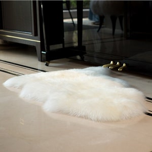 May include: A white fluffy sheepskin rug with long, soft fur. The rug is laid out on a light-colored floor.