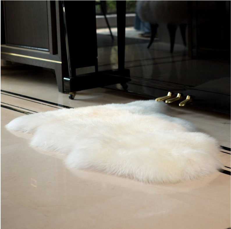 May include: A white fluffy sheepskin rug with long, soft fur. The rug is laid out on a light-colored floor.