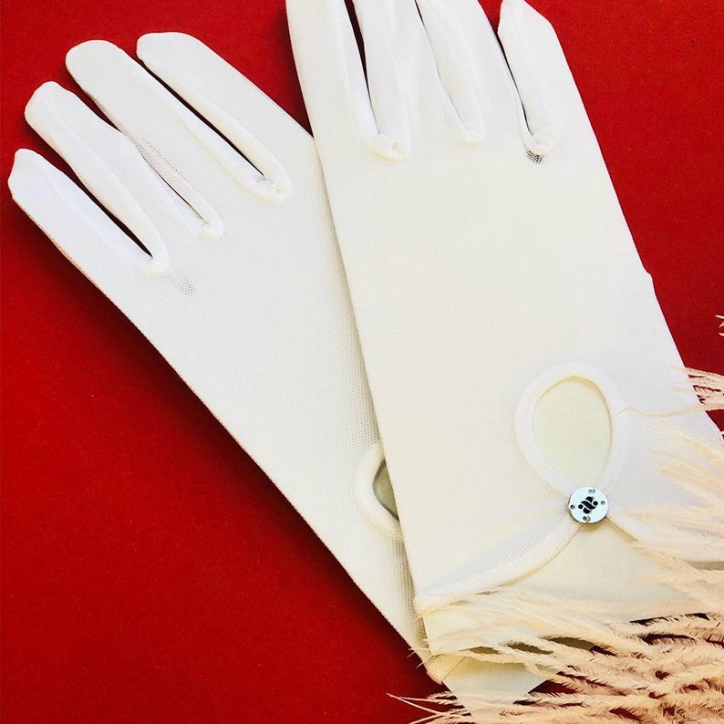 Puede incluir: Un par de guantes blancos con ribete de plumas. Los guantes tienen un peque&ntilde;o bot&oacute;n en la mu&ntilde;eca.