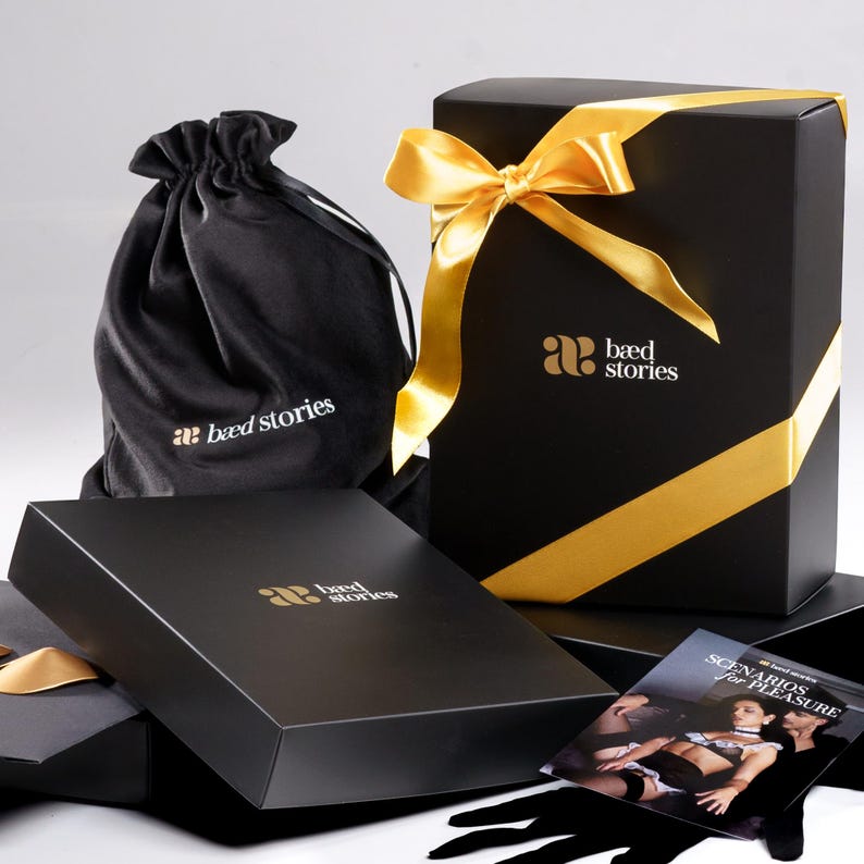 Puede incluir: Una caja de regalo negra con una cinta dorada y una bolsa de terciopelo negro. La caja tiene el texto "bed stories" impreso en ella. La bolsa tiene el texto "bed stories" impreso en ella. La caja est&aacute; sentada sobre una superficie negra.